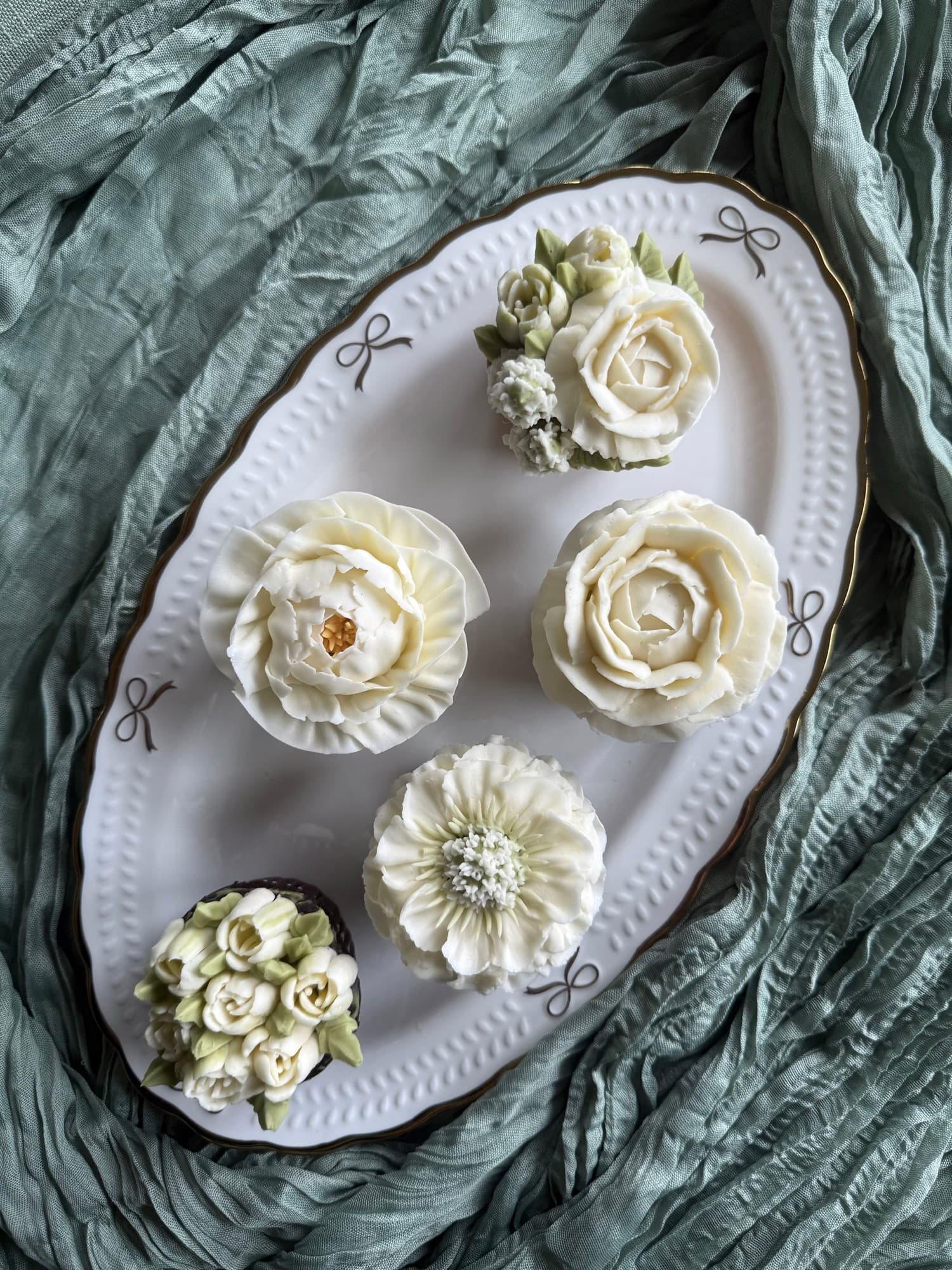Ivory buttercream roses and flower cupcakes on a white platter over sage fabric