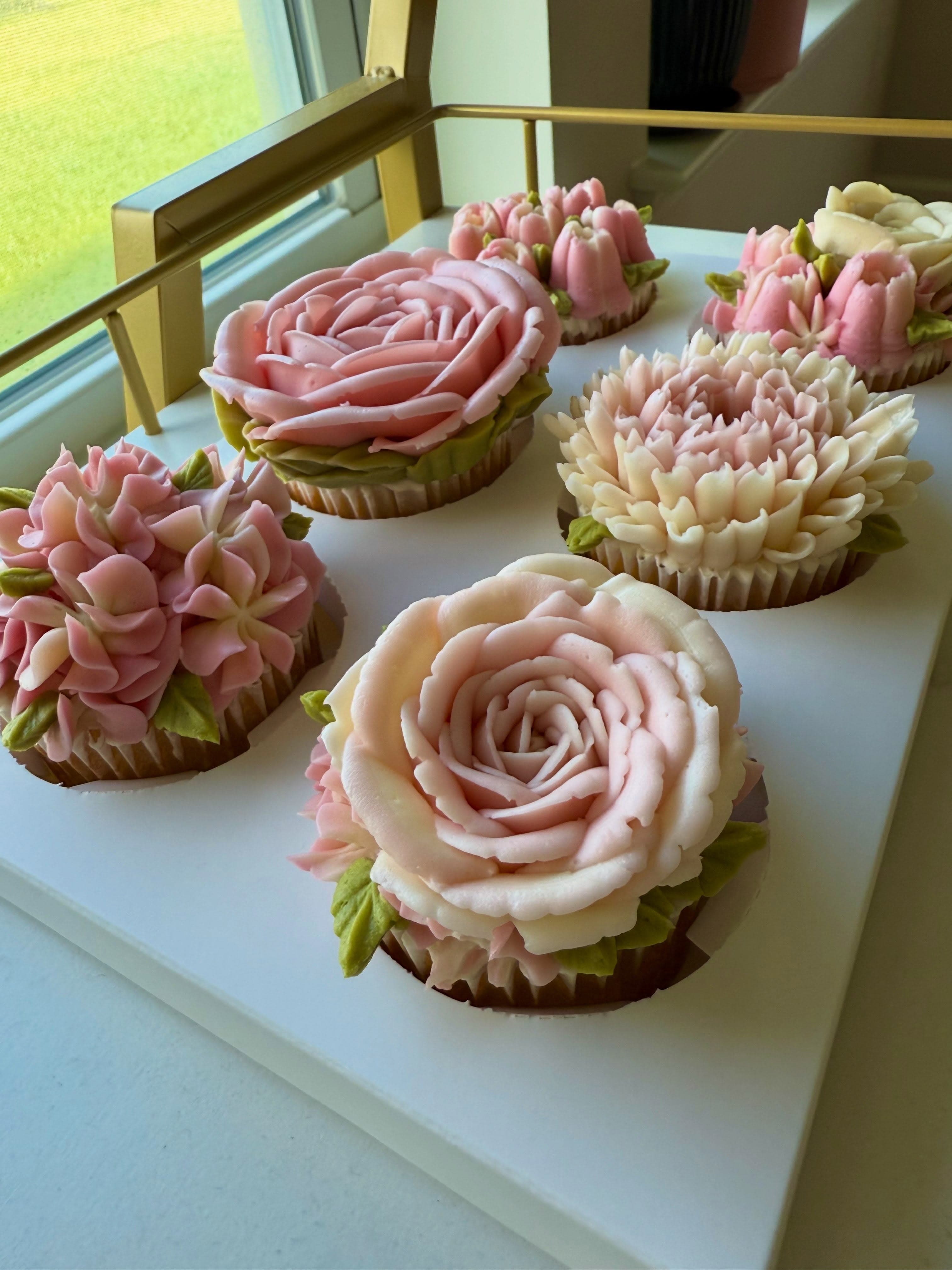 Six cupcakes piped with buttercream roses, hydrangeas and chrysanthemums on a tray in window light