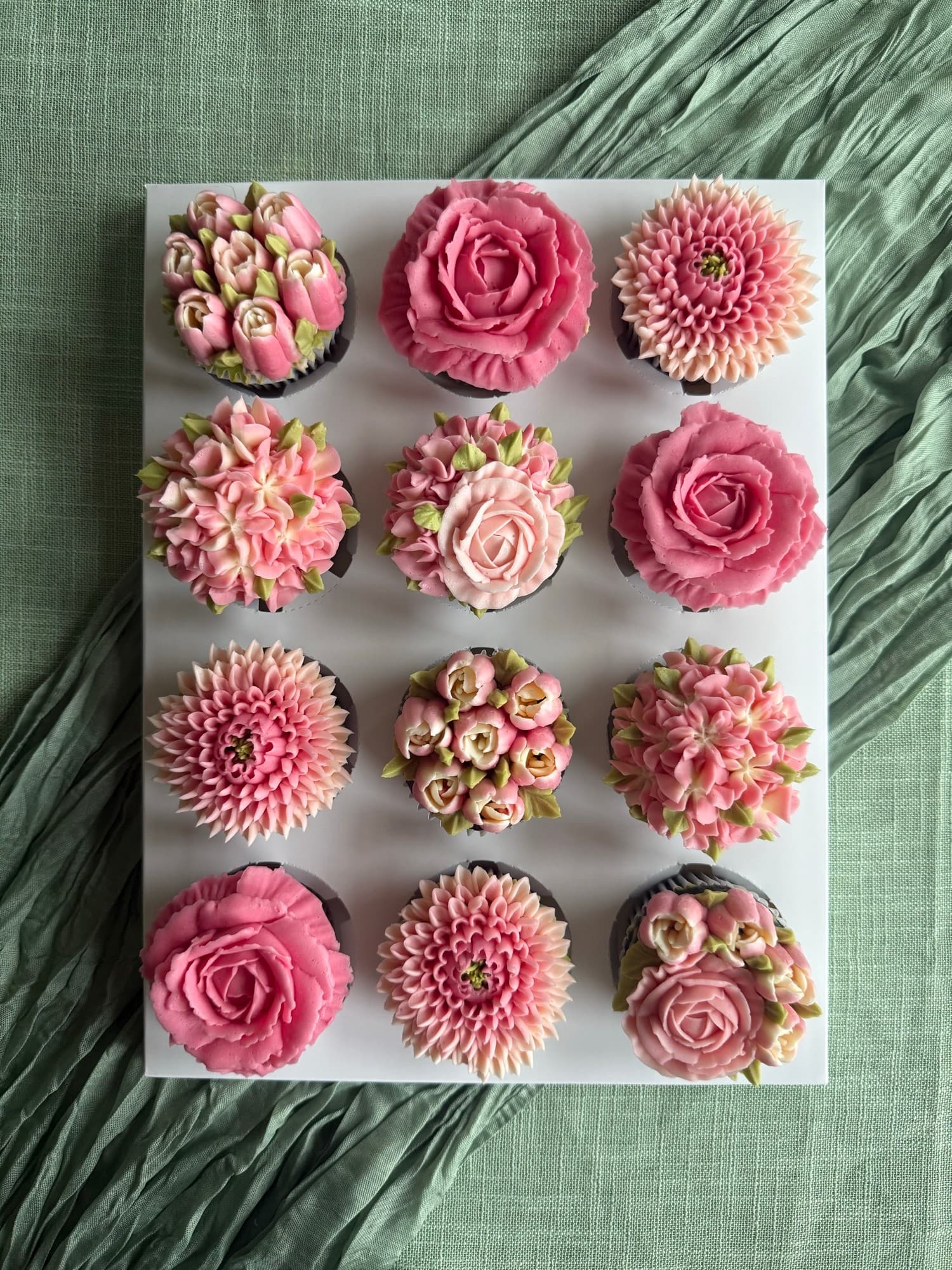 A dozen pink buttercream roses, chrysanthemums and tulips on a white board over sage fabric