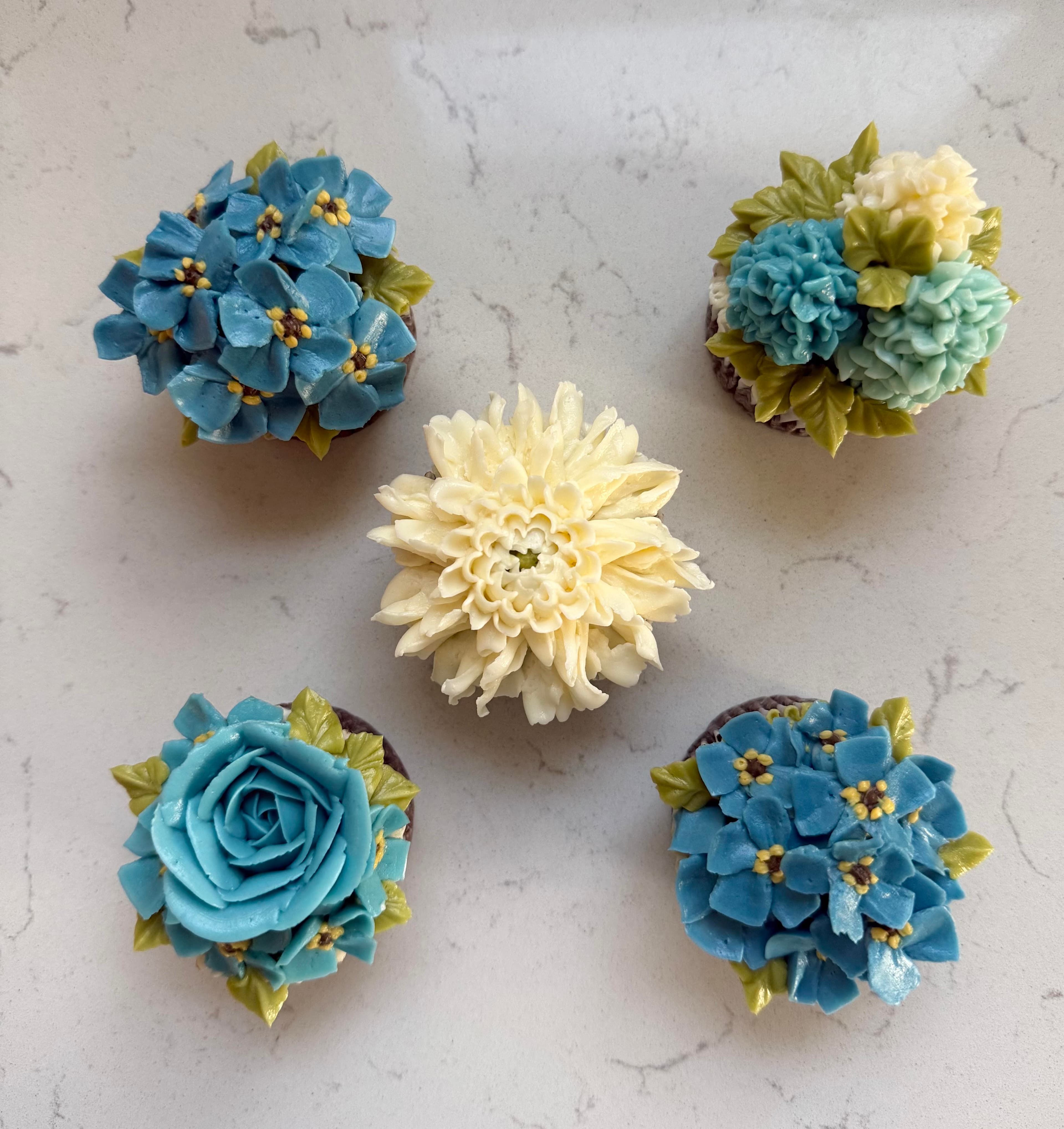 Five cupcakes in blues on marble: hydrangeas, a teal rose and an ivory dahlia
